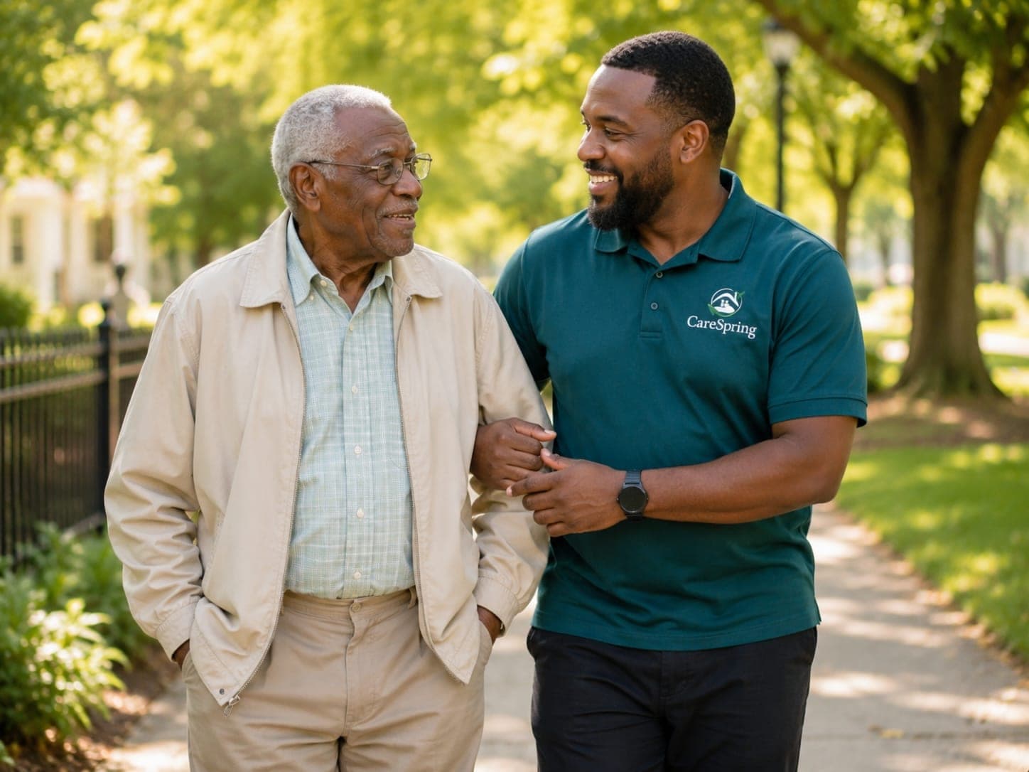Two people walking together outdoors on a sunny path, one in a teal polo shirt with a CareSpring logo offering arm support